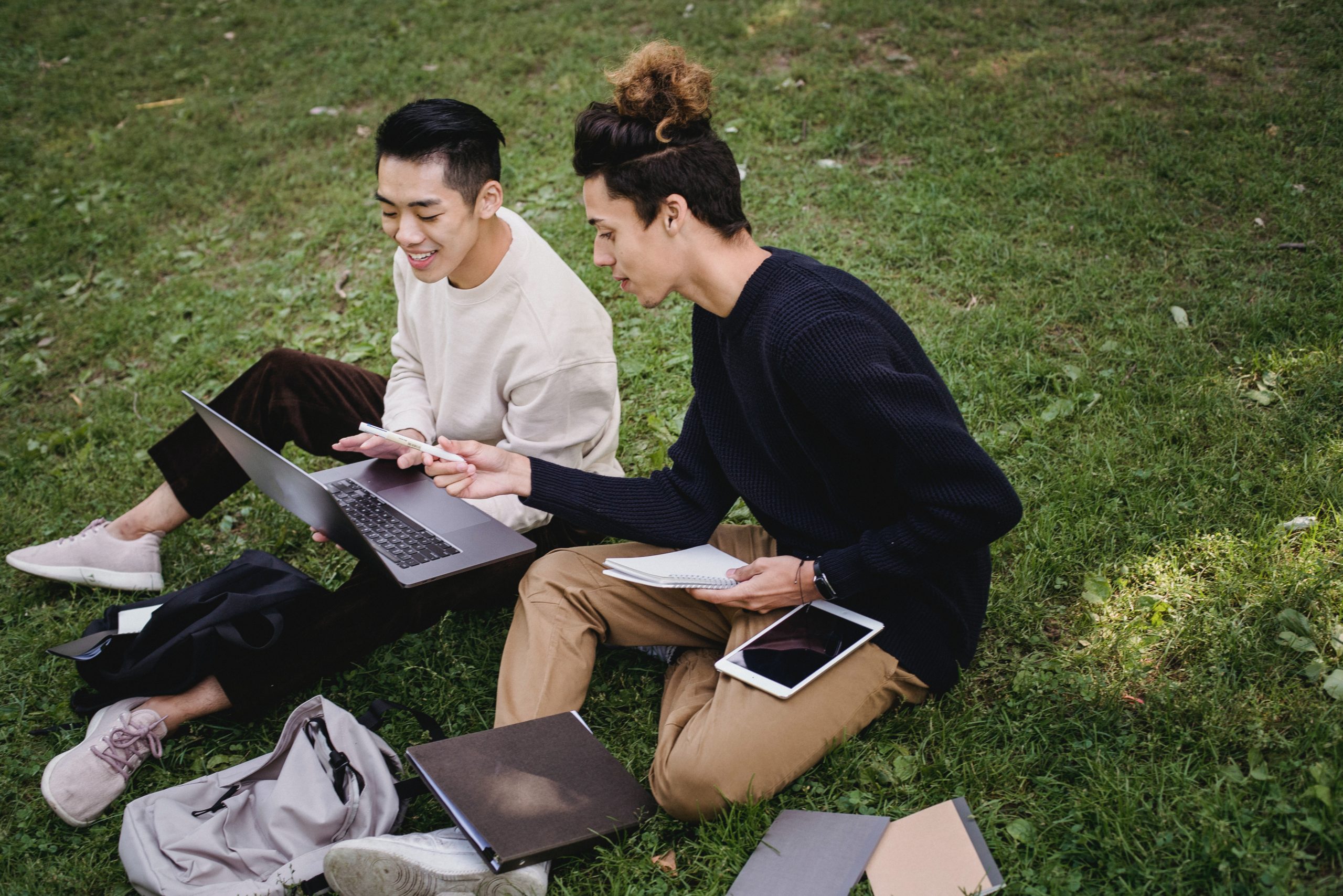 Two students sitting outside on the grass with their books and laptops open.